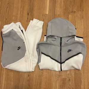 Grå och vit huvtröja från Nike - Säljer ett snyggt grå och vit set från Nike. Tröjan har en stilren design med svarta detaljer och en ficka på ärmen. Till tröjan har vi även ett matchande par byxor. Perfekt för en sportig look eller avslappnade dagar. 