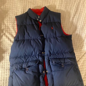 Vändbar dunväst Ralph Lauren - Snygg vändbar dunväst från Ralph Lauren i blått och rött. Västen har knappar och två fickor framtill. Perfekt för att hålla värmen med stil. Den är utmärkt skick. Passar även M. Priset kan diskuteras. 