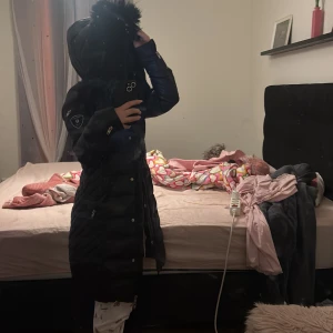 Svart dunjacka från Rockandblue - Säljer en svart lång dunjacka från Rockandblue med pälskantad huva. Jackan har dragkedja och knappar framtill samt en snygg logga på ärmen. Perfekt för kalla dagar! 🖤köptes för 3500
