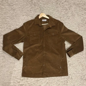 Brun manchesterjacka från Barbour - Säljer en snygg brun manchesterjacka från Barbour i storlek L. Jackan har en klassisk design med knappar framtill och två bröstfickor med lock. Perfekt för höst och vår med sin stilrena look och bekväma passform.