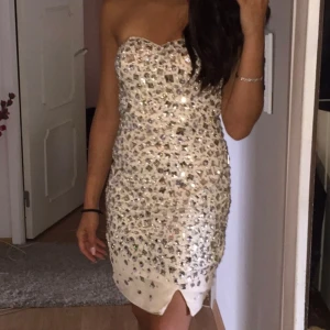 Glittrig axelbandslös klänning - Säljer en superfin axelbandslös klänning i beige med massor av glittriga paljetter. Perfekt för en festlig kväll! Klänningen har en tight passform och en kort längd som ger en elegant look.