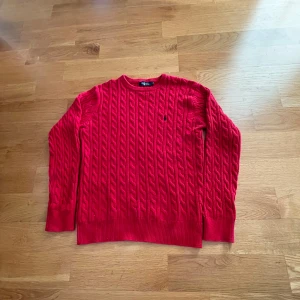 Röd stickad tröja från Ralph Lauren - Säljer en snygg röd stickad tröja från Ralph Lauren med kabelstickat mönster och broderad logga på bröstet. Perfekt för kyligare dagar med sin klassiska design och bekväma passform. Tröjan är använd ungefär 10 gånger. Säljer för att den är förstor. 