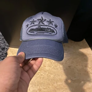 Blå keps med stjärnor - Snygg blå keps med broderade stjärnor och ett motiv framtill. Kepsen har ett nätmaterial på sidorna och justerbar passform. Perfekt för en avslappnad stil.