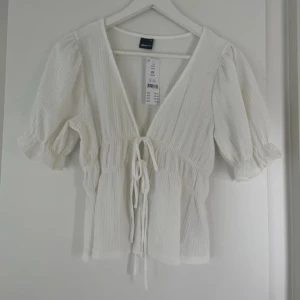 Oanvänd Vit Daisy topp / blus från Gina tricot  - Säljer en söt vit Daisy topp med v-ringning och knytning framtill. Helt ny o oanvänd. Toppen har korta puffärmar och en lätt ribbad struktur som ger en fin textur. Perfekt för en somrig look!
