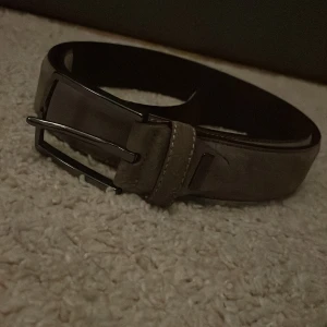 Grått mocka bälte  med metallspänne - Snyggt grått mocka bälte med ett stilrent metallspänne. Perfekt för att ge en klassisk touch till din outfit. Skärpet har flera hål för justerbar passform. Bältet kostar 600 kr nypris, strl 85 cm 