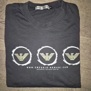 Grå långärmad t-shirt från Emporio Armani storlek 158/164 - Snygg grå långärmad t-shirt från Emporio Armani med tre ikoniska logotyper tryckta på framsidan. Perfekt för en stilren och avslappnad look. Tillverkad i mjukt material för extra komfort. Storlek 158/164 cl