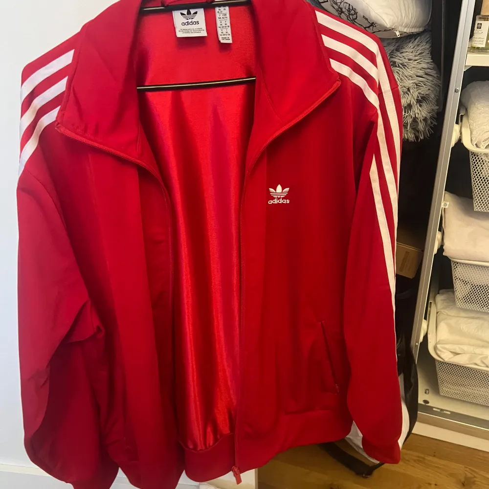 Säljer en klassisk röd träningsjacka från Adidas med vita ränder längs ärmarna. Jackan har dragkedja framtill och två fickor. Perfekt för en sportig look eller avslappnad stil. 🏃♂️. Takit.