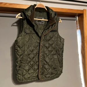  Quiltad väst med luva - Snygg grön quiltad väst från Sätila med dragkedja och knappar. Perfekt för lager-på-lager under höst och vår. Västen har praktiska fickor och en bekväm passform. Saknar en knapp längst ner, men de är inget som påverkar