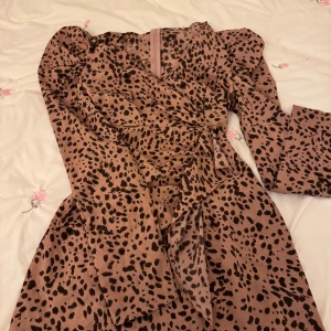 Mönstrad omlottklänning - Säljer en snygg omlottklänning med leopardmönster i bruna toner. Klänningen har långa ärmar och en knytning i midjan för en smickrande passform. Perfekt för en trendig look!