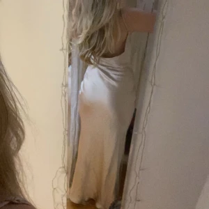 Champagnefärgad balklänning - Elegant champagnefärgad långklänning med smala axelband och en mjuk, flytande siluett. Klänningen har en vacker drapering framtill och en öppen rygg som ger en sofistikerad look. Perfekt för speciella tillfällen.