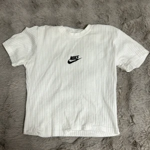 Vit ribbad t-shirt från Nike - Säljer en stilren vit ribbad t-shirt från Nike med korta ärmar och en klassisk Nike-logga på bröstet. Perfekt för en avslappnad look. Den är inte äkta 