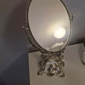Vintage smyckespegel - Vacker ovalt smyckespegel med detaljerad silverram och dekorativa blommönster. Spegeln står på en utsmyckad fot som ger en klassisk och lyxig känsla. Perfekt för att ge en elegant touch till ditt rum. Fick den vintage spegeln av min mormor