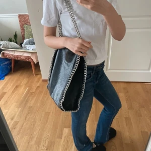 TIAMO Svart axelväska med kedjedetaljer liknande till stella mccartney - Snygg svart axelväska med silverfärgade kedjedetaljer längs kanterna och axelremmen. Väskan har en dragkedja på baksidan för extra förvaring. Perfekt för att ge en edgy touch till din outfit.