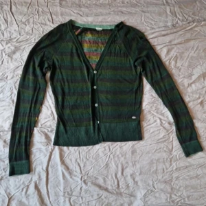 Grönrandig kofta från Skunkfunk - Säljer en grönrandig kofta från Skunkfunk med knappar framtill. Koftan har långa ärmar och en v-ringad design. Perfekt för lager-på-lager-stil och tillför en färgglad touch till din garderob.