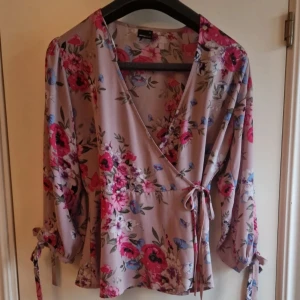 Blommig omlottblus från Gina Tricot - Säljer en vacker blommig omlottblus från Gina Tricot i storlek 38. Blusen har ett färgglatt blommönster i rosa, blått och rött på en beige bakgrund. Den har trekvartsärmar med knytband och en v-ringad omlottdesign som ger en elegant touch.