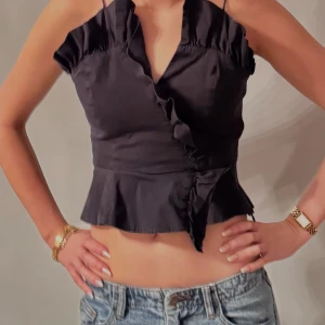 Svart fest topp  - Snygg svart omlottlinne med volangdetaljer och knytning i midjan. Perfekt för en stilren look. Axelbandslös design som ger en elegant touch.