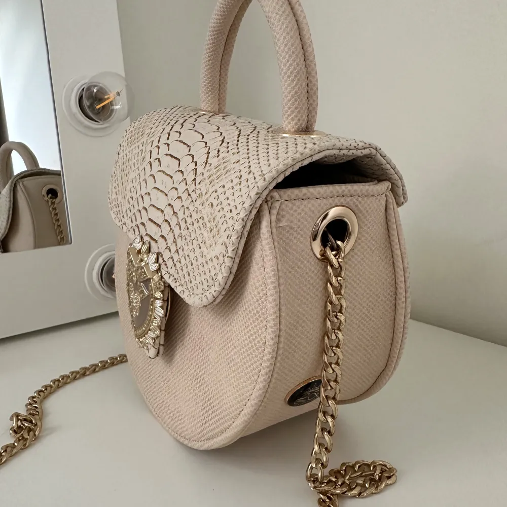 Säljer en superfin beige rund handväska med en lyxig guldkedja. Väskan har ett snyggt ormskinnsmönster på locket och en dekorativ hjärtformad detalj framtill. Perfekt för att ge din outfit en elegant touch! Väskan är köpt utomlands för några år sen🙂. Väskor.