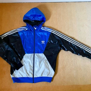 Adidas vindjacka stl L - Klassisk Adidas jacka i glansigt material, bra skick med endast ett litet hål - se bilder. Huva och fickor med dragkedja.