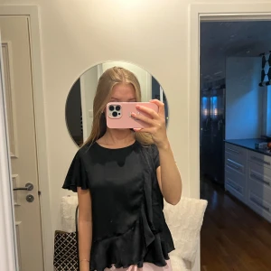 Svart blus med volangdetaljer - Säljer en elegant svart blus med korta ärmar och volangdetaljer vid nederkanten. Perfekt för både vardag och festliga tillfällen. Blusen har en lös passform som ger en bekväm känsla.