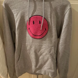 Grå hoodie från Sandro med smiley - Säljer en grå hoodie från Sandro med en stor rosa smiley på framsidan. Tröjan har en klassisk passform med långa ärmar och en bekväm huva med dragsko. Perfekt för en avslappnad stil! Använd fåtal gånger