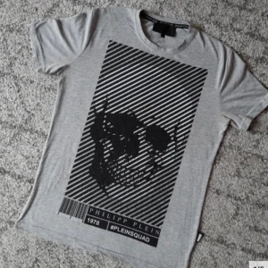 Grå t-shirt med dödskalle från Philipp Plein - Snygg grå t-shirt från Philipp Plein med ett stort svart dödskalletryck på framsidan och textdetaljer på baksidan. Tillverkad i 100% bomull för en bekväm känsla. Perfekt för en cool och avslappnad stil. Nypris 2500kr. Storleken är L men sitter snarare som M. Original givetvis! Passa på att fynda.