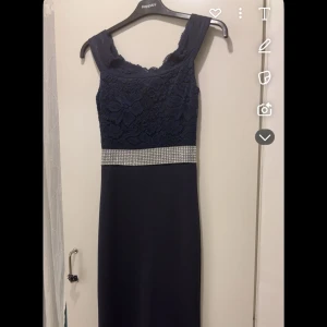 Mörkblå långklänning med spets och strassdetaljer - Elegant mörkblå långklänning med spetsöverdel och strassbälte i midjan. Klänningen har en figurnära passform och är perfekt för formella tillfällen. Har även en kort slits bakifrån, original pris 4000kr och priset kan även diskuteras klänningen är även off shoulder det syns bara inte för att den hänger❤️