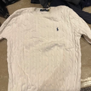 Vit stickad tröja från Ralph Lauren - Säljer en klassisk vit stickad tröja från Ralph Lauren med kabelstickat mönster. Tröjan har en broderad logotyp på bröstet och långa ärmar. Perfekt för en stilren och tidlös look.