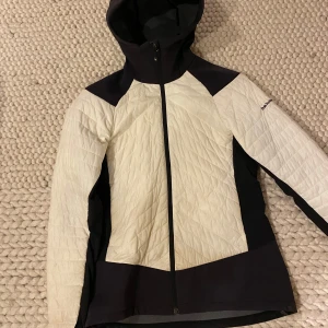 Vit och svart jacka från Peak Performance - Säljer en snygg vit och svart jacka från Peak Performance. Jackan har en dragkedja framtill och en praktisk huva. Perfekt för kyliga dagar med sin stilrena design. Höst/vår jacka