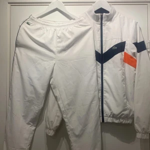 Vit vindjacka och byxor från Lacoste - Säljer en snygg vit vindjacka och vindbyxor från Lacoste med coola marinblå och orange detaljer. Jackan har en dragkedja framtill och elastiska muddar vid ärmslut och nederkant. Perfekt för träning 🧡💙