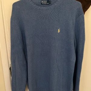 Blå tröja från Polo Ralph Lauren - Säljer en stilren blå tröja från Polo Ralph Lauren med det klassiska broderade logotypen i gult på bröstet. Tröjan är långärmad och har en ribbad krage samt muddar vid ärmslut och nederkant. Perfekt för en avslappnad och snygg look. Den har inga flaws eller tecken på användning, men är en äldre modell! Står storlek: M, men sitter som L