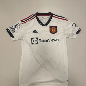 Vit Manchester United fotbollströja - Säljer en vit Manchester United fotbollströja från Adidas med korta ärmar. Tröjan har klubbens emblem på bröstet och 'Rashford 10' tryckt på ryggen. Den har svarta och röda detaljer samt sponsorer som TeamViewer och DXC Technology. Denna united tröjan är köpt i old trafford i England ” Manchester united hemma arena” tröjan har inga defekter men bara att etiketten vid höften är bort klippt. Nypris 1500. Kontakta mig vid minsta fråga👍🏼