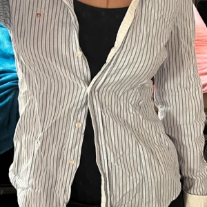 Randig skjorta från GANT - Säljer en stilren och klassisk vit och blå randig skjorta från GANT. Skjortan har långa ärmar och knappar framtill. Perfekt för en elegant och avslappnad look.