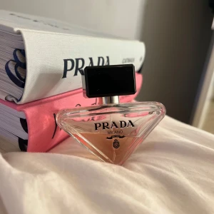 Prada Milano parfym - Elegant parfym från Prada Milano i en stilren, triangulär glasflaska med svart fyrkantigt lock. Flaskan har en ljusrosa nyans som ger en lyxig känsla. Perfekt för den som vill ha en sofistikerad doftupplevelse. Mer än halva kvar