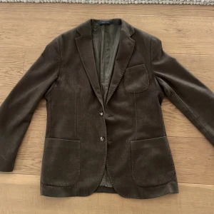 Brun kavaj i italienskt tyg - Snygg brun kavaj med två knappar och fickor fram. Tillverkad av fint italienskt tyg, perfekt för en stilren look. Kavajen har en klassisk design med knappdetaljer vid ärmslut och en bröstficka.