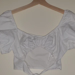 Vit kortärmad crop top - Säljer en söt vit blus med korta puffärmar och knytband framtill. Blusen har en öppen design vid midjan och är perfekt för sommardagar. Den är gjord i ett lätt och bekvämt material.