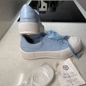 Blå sneakers från Alexander McQueen - Snygga ljusblå sneakers från Alexander McQueen med vit sula och matchande blå snörning. Skorna har en stilren design med märkets logotyp diskret placerad på hälen. Perfekta för en trendig och avslappnad look. Två skosnören tillkommer de vita är helt oanvända som kom till när jag köpte dem, de blåa är använda. PRIS KAN DISKUTERAS