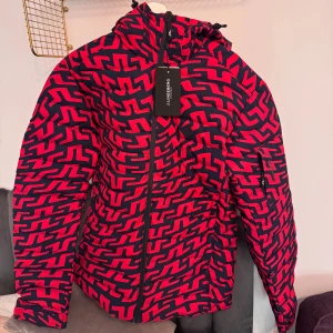 Röd och svart mönstrad skidjacka från J.Lindberg - Snygg och färgglad jacka från J.lindberg i rött och svart med ett unikt mönster. Jackan har en dragkedja framtill och en praktisk huva. Perfekt för att sticka ut i mängden! Helt oanvänd med prislapp, säljer även matchande skidbyxor 