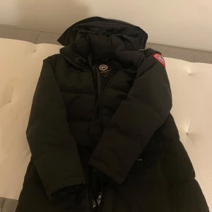 Svart dunjacka från Canada Goose - Säljer en svart dunjacka från Canada Goose med huva och dragkedja. Perfekt för kalla vinterdagar. Jackan har en klassisk design med lång ärm och är tillverkad i slitstarkt material. Dragkedjan är trasig där av priset