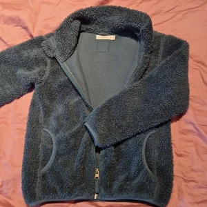 Blå fleecejacka från Minimarket - Mysig blå fleecejacka från Minimarket med dragkedja framtill och två fickor. Perfekt för kyliga dagar med sin mjuka och varma design. Jackan har en hög krage för extra värme.