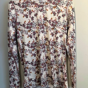 Långärmad tröja från Kari Traa - Säljer en snygg långärmad tröja från Kari Traa med ett blommigt mönster i rosa, orange och svart på en vit bakgrund. Tröjan är tillverkad i en mjuk polyesterblandning och har en normal passform. Perfekt för vår och sommar!