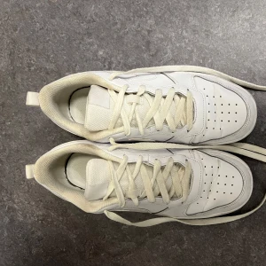 Vita Nike sneakers - Säljer ett par vita Nike sneakers med klassisk design och snörning. Skorna har en stilren look med perforerade detaljer för ventilation. Perfekta för en avslappnad stil.