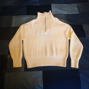 Vit ribbstickad tröja i bommul från Axel Arigato - Säljer en stilren vit ribbstickad tröja från Axel Arigato med dragkedja och hög krage. Tröjan har långa ärmar och ett tryck på ryggen med märkesnamnet. Nypris 2700 kr. Perfekt för en trendig look!