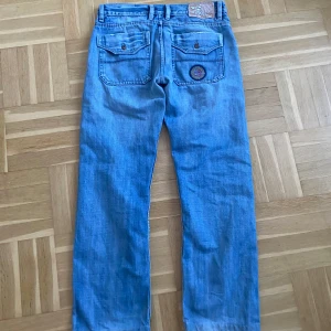 Blå jeans från Diesel - Snygga blå jeans från Diesel med raka ben och klassisk femficksdesign. Byxorna har en knappdetalj på bakfickorna och en broderad patch. Perfekta för en avslappnad stil. Står storlek 32 och passar mig som har s/m. PRISET GÅR ALLTID ATT DISKUTERAS🥰🥰