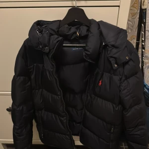 Ralph lauren jacka  - Säljer en mörkblå pufferjacka från Polo Ralph Lauren med huva som är av tag bar. Jackan är sjukt snygg och skön knappt använd också. och är perfekt för kyliga dagar. Den är både stilren och varm. Köpt från zalando nypris hela 5500kr