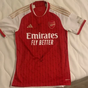 Singnerad Röd Arsenal fotbollströja med Martinelli 11 - Säljer en röd Arsenal fotbollströja från Adidas med Martinelli 11 på ryggen. Tröjan har korta vita ärmar och ett subtilt randigt mönster. Den är prydd med sponsorloggan 'Emirates Fly Better' på framsidan och har guldiga Adidas-ränder på axlarna. Den är även singnerad av Arsenal spelarna Trossard och Martinelli