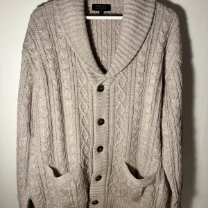 Beige stickad kofta med knappar - Säljer en mysig beige stickad kofta från indigo. Koftan har ett klassiskt flätmönster och stora knappar framtill. Den har långa ärmar och två praktiska fickor. Perfekt för kyliga dagar! Aldrig använd