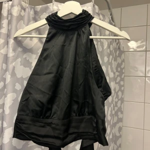 Svart halterneck topp - Elegant svart halterneck topp med knytning i nacken och en dekorativ rosett framtill. Perfekt för en stilren look.
