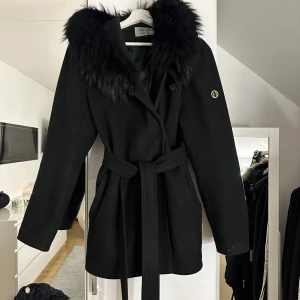 Svart kappa från Hollies - Elegant svart kappa från Hollies med en lyxig pälsdetalj på huvan och ett knytskärp i midjan.  Använd cirka en höst, den är super bra i storleken passar även någon som har S/M, M/L. Fint skick. Storlek 44