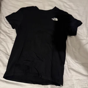 Svart t-shirt från The North Face - Snygg svart t-shirt från The North Face no med logga på bröstet och stor röd logga med texten 'Never Stop Exploring' på ryggen. Perfekt för en avslappnad stil.