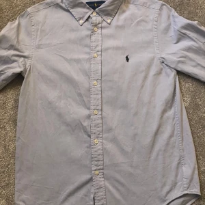 Ljusblå skjorta från Ralph Lauren - Säljer en klassisk ljusblå skjorta från Ralph Lauren med knappar och broderad logga på bröstet. Skjortan har en stilren design med långärmade ärmar och en bekväm passform.